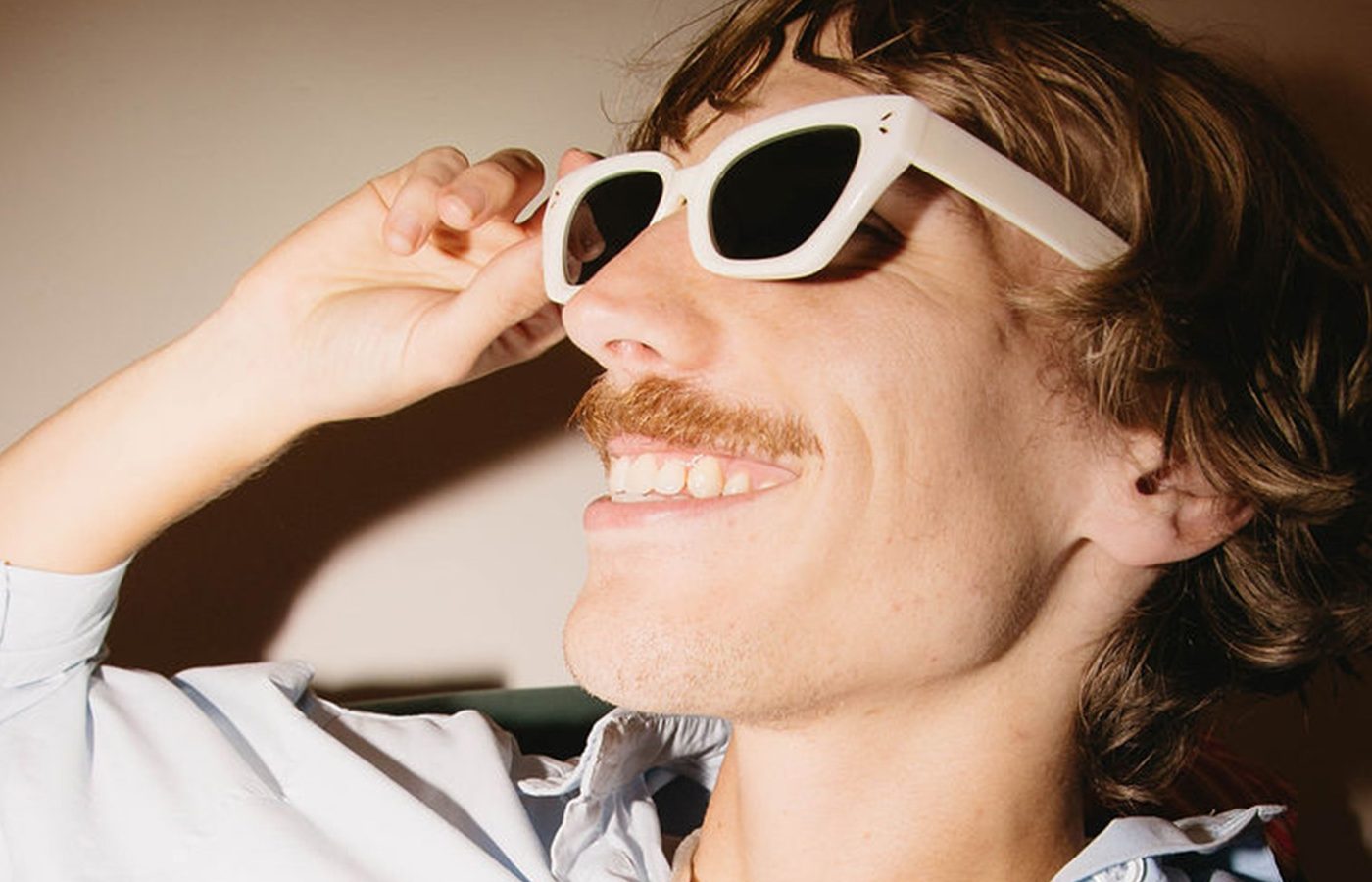 A smiling man with a mustache adjusts thick white sunglasses while wearing a light-colored shirt.