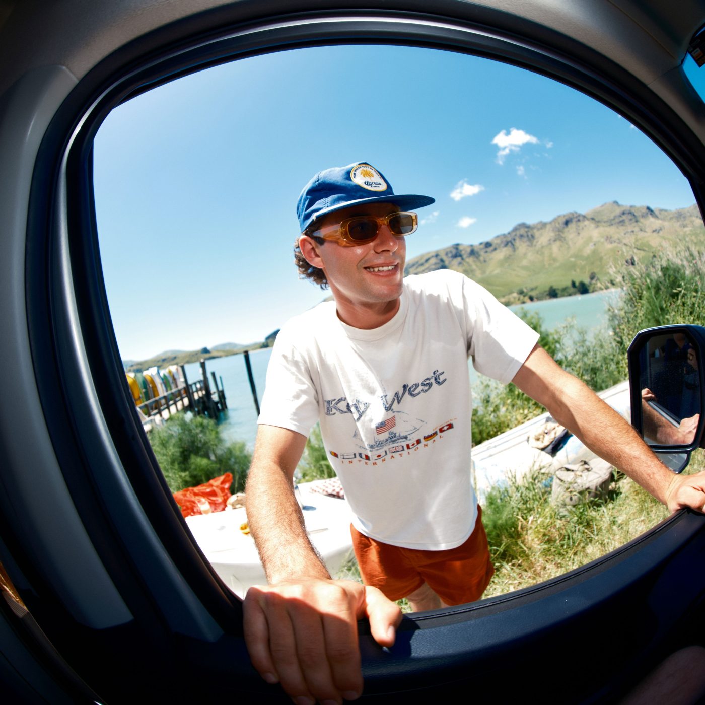 Smiling man in a blue cap and sunglasses leans into a car window beside a lake and mountains.