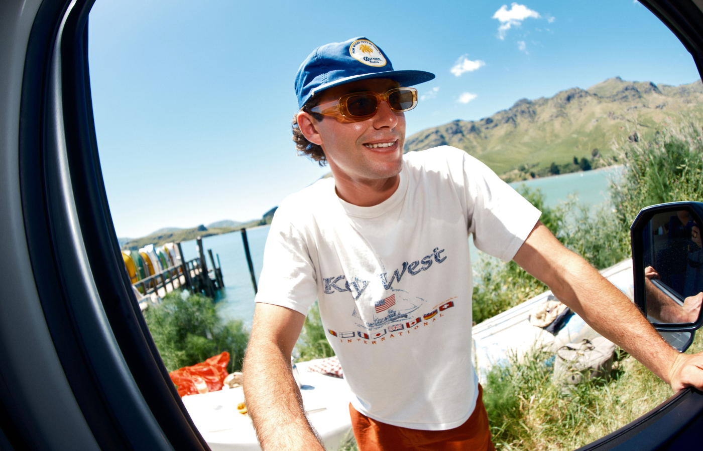 A smiling man in a blue cap and sunglasses leans into an open vehicle beside a lake and mountains.