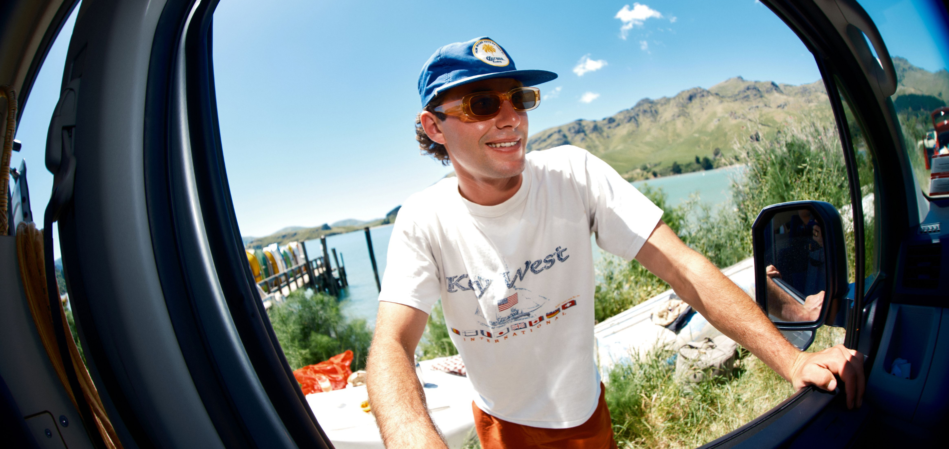 A smiling man in a blue cap and sunglasses leans into an open vehicle beside a lake and mountains.