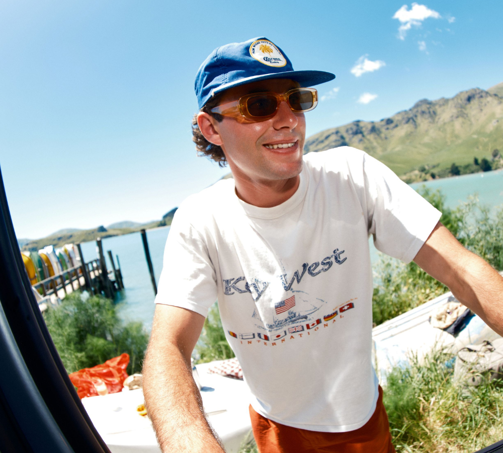 A smiling man in a blue cap and sunglasses leans into a vehicle beside a lake, with mountains in the background.