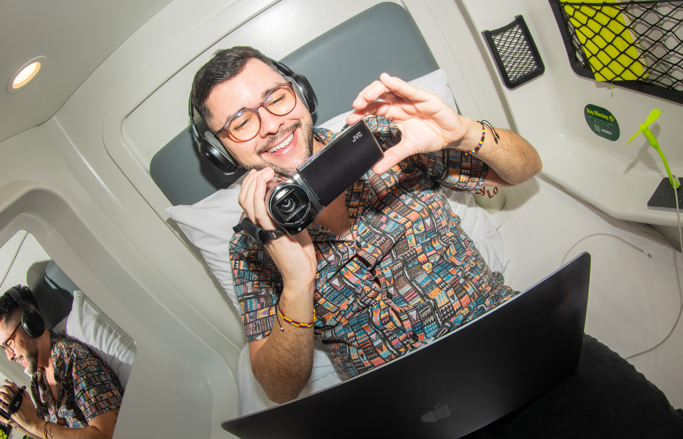 Smiling man wearing headphones reclines in a compact pod, holding a camcorder above an open laptop; his reflection appears in a mirror.