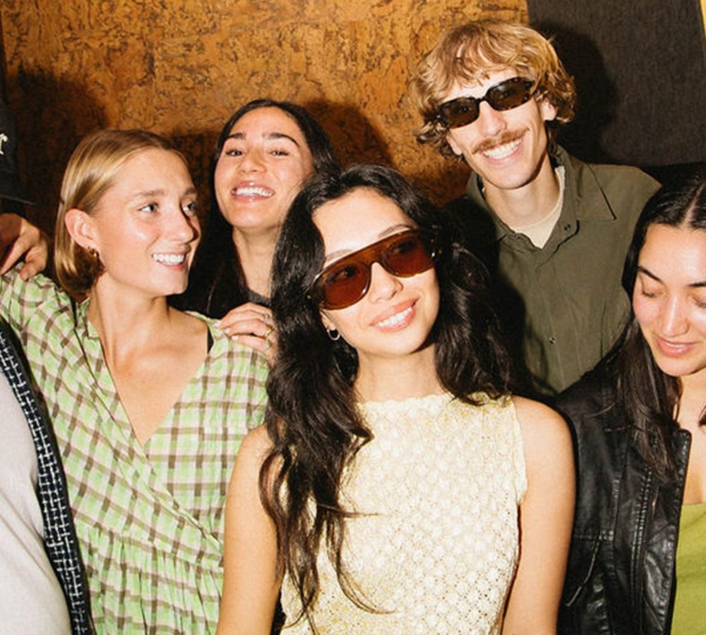 Six people pose together indoors; a man in a cap flashes a peace sign while others smile, some wearing sunglasses.